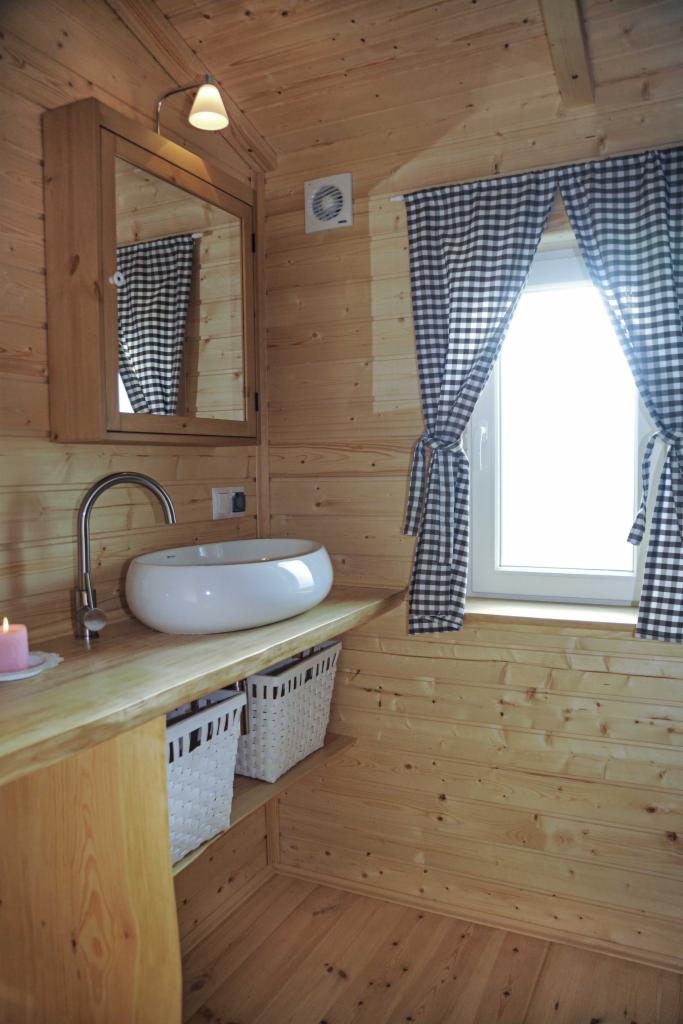 Drewniana łazienka z umywalką nablatową, lustrem w drewnianej ramie i oknem z zasłonami w czarno-białą kratę. Pod blatem umywalki widoczne białe wiklinowe kosze.