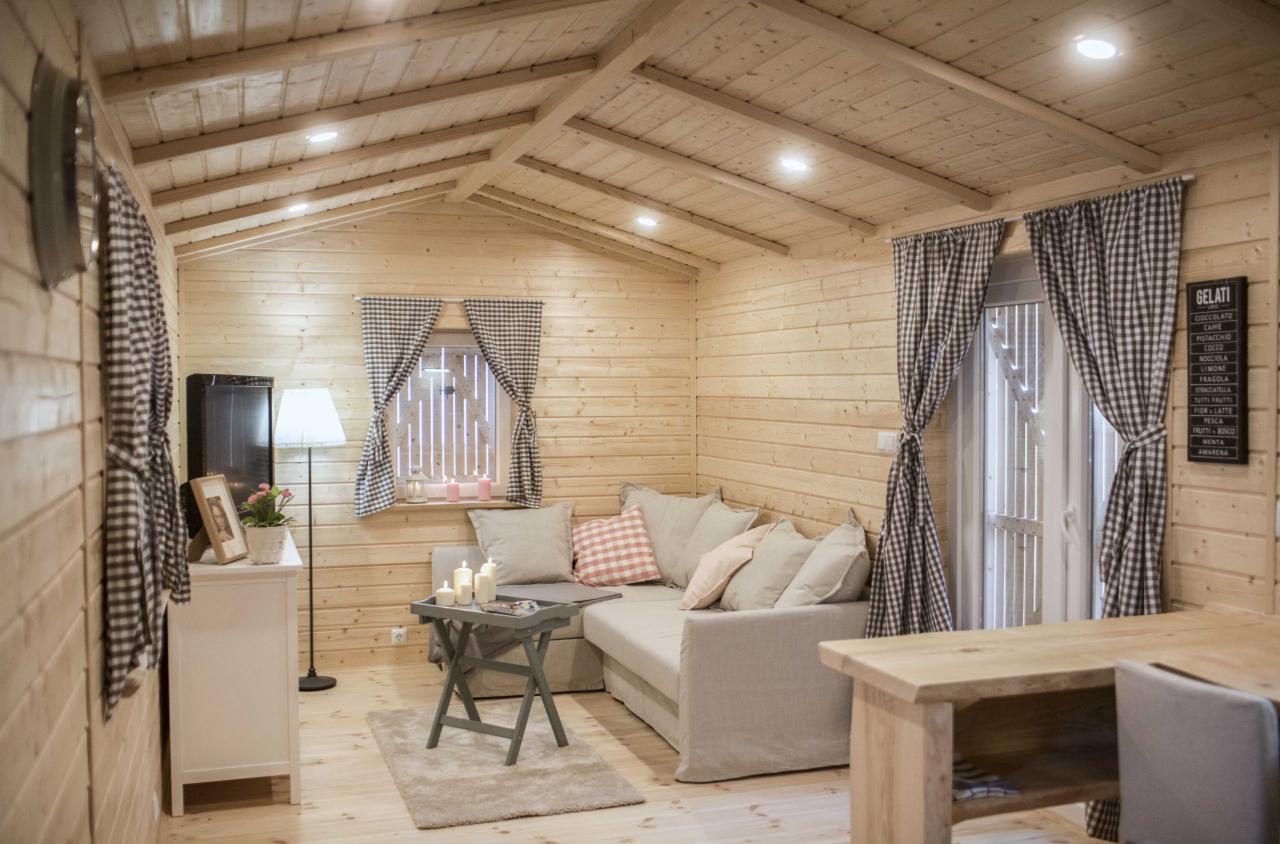 Jasne, drewniane wnętrze salonu z narożną sofą, stolikiem kawowym ze świecami, oknem z zasłonami w kratę i drewnianym stołem w prawym dolnym rogu.