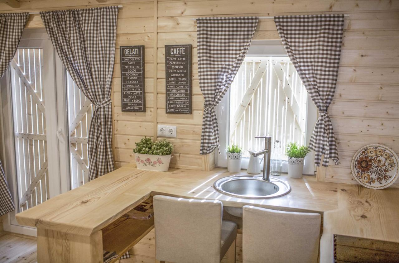 Jasna, drewniana kuchnia w domu z bali, z oknem z drewnianymi okiennicami i zasłonami w kratę, drewnianym blatem, zlewozmywakiem i dekoracjami w postaci doniczek z ziołami i talerza z motywem...