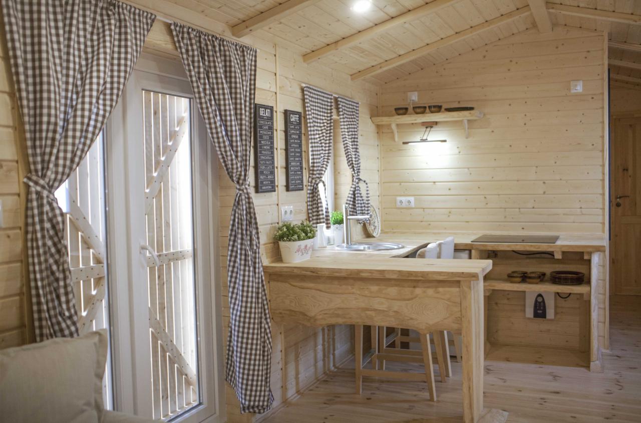 Wnętrze drewnianego domu z aneksem kuchennym, widoczne okno z kraciastymi zasłonami i drewniane meble kuchenne.