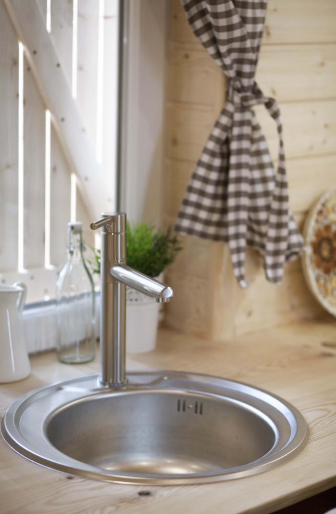 Kuchenny zlew ze stali nierdzewnej z nowoczesną baterią na drewnianym blacie, w tle okno z białymi okiennicami i zasłona w brązowo-białą kratę, wnętrze wykończone drewnem.