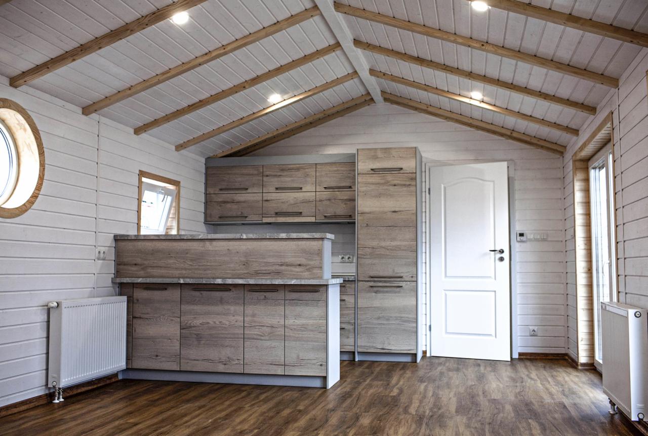 Jasne wnętrze pomieszczenia w stylu skandynawskim z drewnianymi elementami, widoczne białe ściany z desek, drewniana podłoga, zabudowa kuchenna w odcieniach szarości, okrągłe okno oraz białe drzwi.