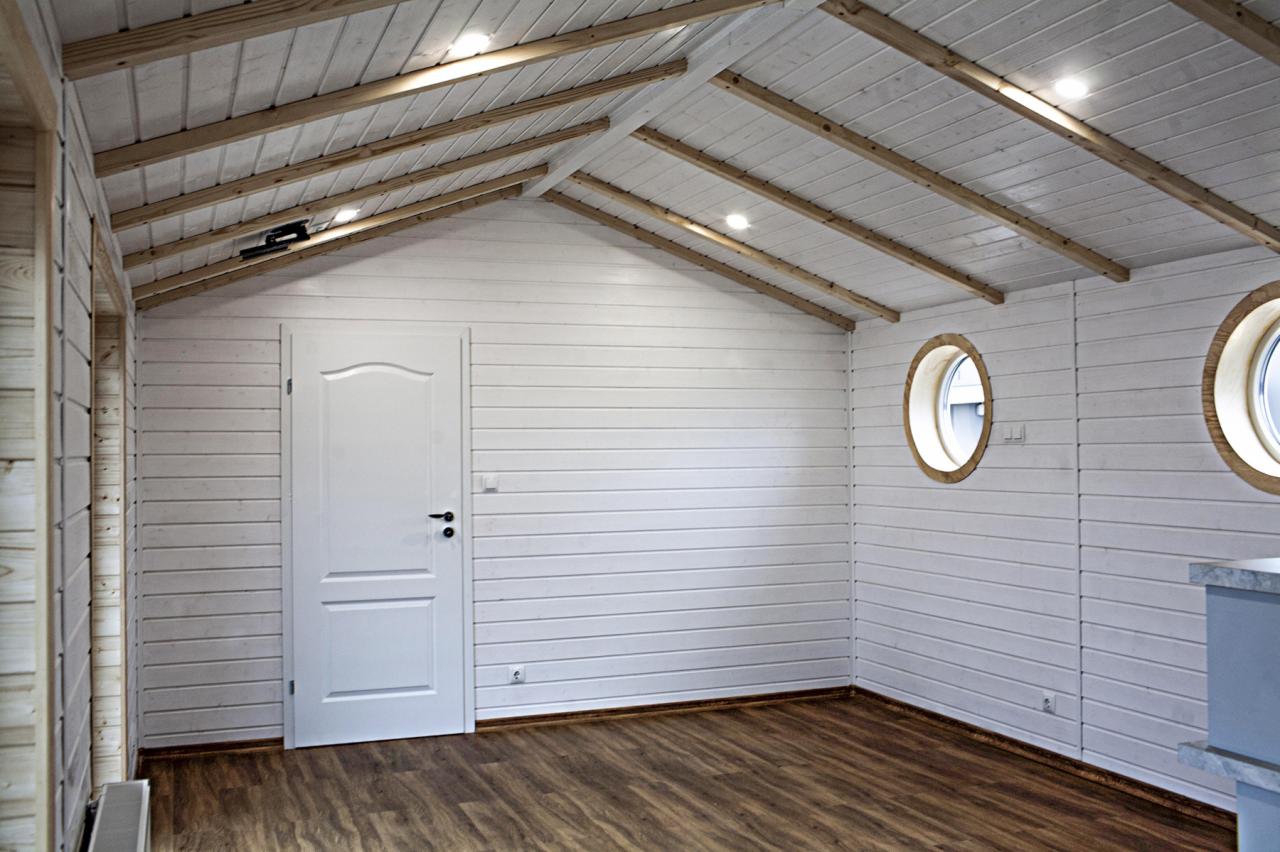 Wnętrze pomieszczenia z drewnianą podłogą i ścianami z białych desek, okrągłe okna z drewnianymi ramami, białe drzwi z czarną klamką, drewniane belki stropowe i punktowe oświetlenie.