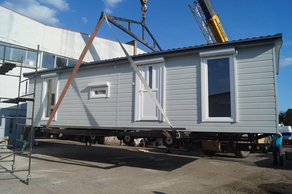 Szary, gotowy moduł mieszkalny z białymi oknami i drzwiami, transportowany przez dźwig na platformie z kołami, na tle błękitnego nieba i budynku przemysłowego.