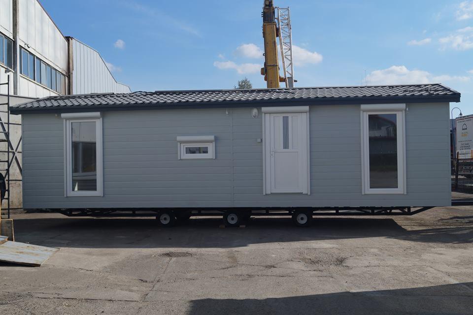 Gotowy, szary, mobilny dom z oknami, drzwiami i roletami zewnętrznymi, ustawiony na betonowym podłożu z dźwigiem w tle.