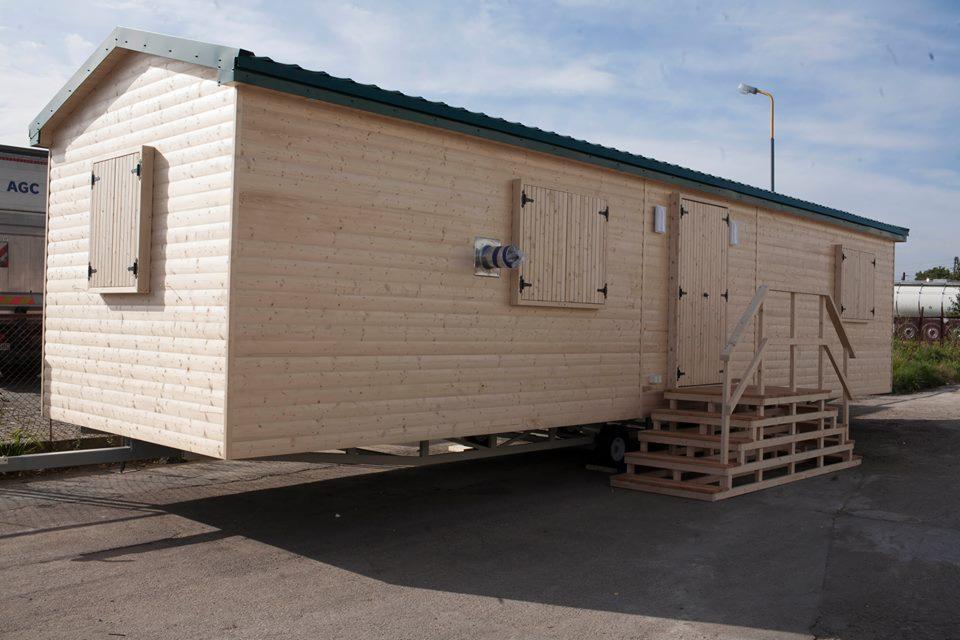 Przenośny, parterowy budynek modułowy z jasnego drewna, posadowiony na stalowej ramie z kołami, z drewnianymi okiennicami, schodami i zielonym dachem