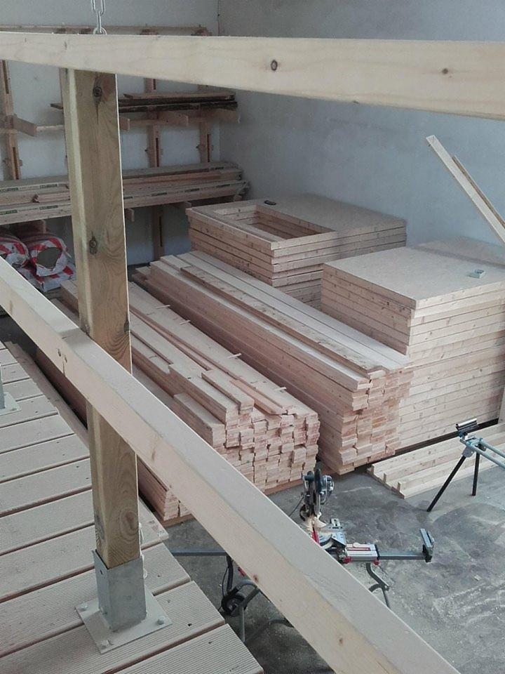 Stosy drewnianych elementów konstrukcyjnych, w tym ramy i płyty, przygotowane do montażu wewnątrz budynku, widoczne narzędzia do obróbki drewna.