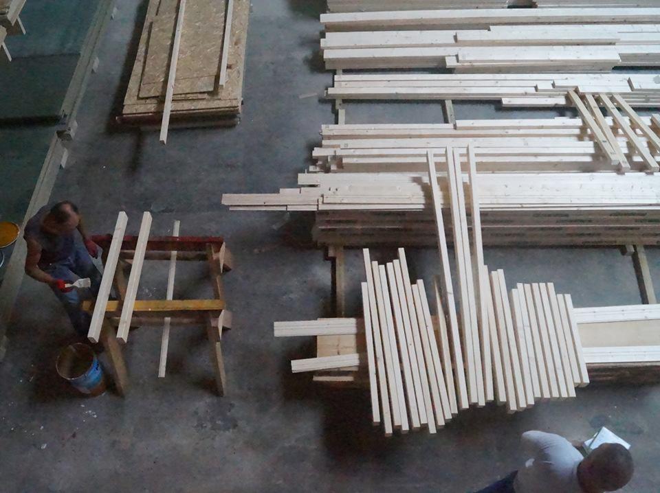 Przygotowanie elementów drewnianych do budowy domu szkieletowego: malowanie belek na kozłach w warsztacie, widoczne stosy nieobrobionego drewna i płyt OSB.