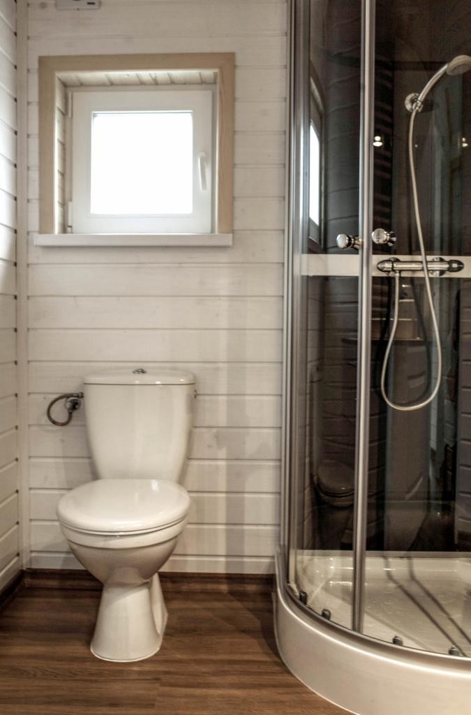 Wnętrze łazienki z kabiną prysznicową i toaletą w domu z białych paneli drewnianych, małe okno z drewnianą ramą, brązowa podłoga.