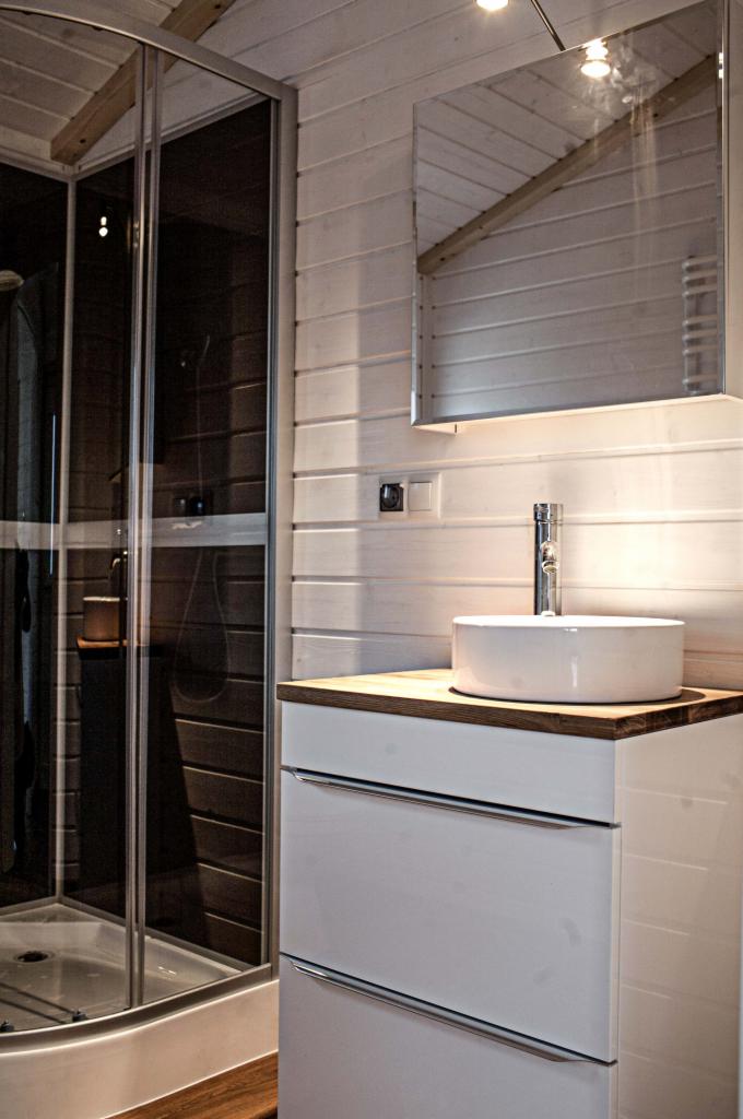 Wnętrze łazienki w domu szkieletowym z kabiną prysznicową, białą umywalką nablatową na szafce z szufladami i lustrem na ścianie z boazerii.