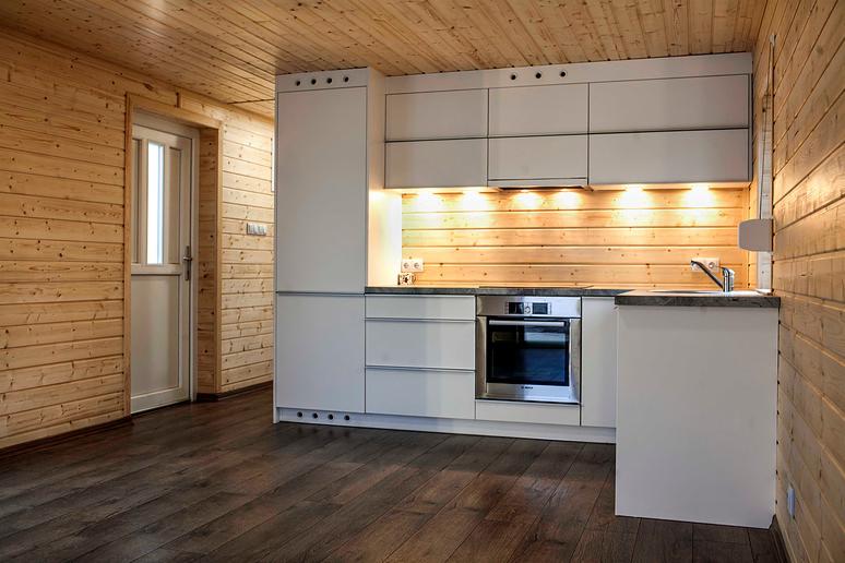 Wnętrze domu szkieletowego z drewnianą boazerią i nowoczesną białą zabudową kuchenną z oświetleniem podszafkowym i sprzętem AGD ze stali nierdzewnej.