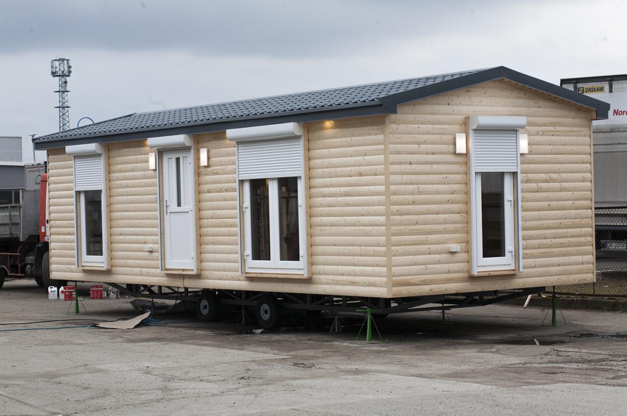 Przenośny modułowy budynek z jasnego drewna, z roletami zewnętrznymi i oświetleniem elewacyjnym, ustawiony na metalowej ramie z kołami i wspornikami, na betonowym placu.