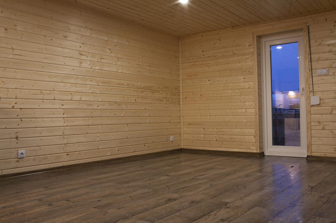 Wnętrze wykończonego pokoju z drewnianymi panelami ściennymi i podłogą imitującą drewno, widoczne gniazdka elektryczne i białe drzwi balkonowe z widokiem na zewnątrz.