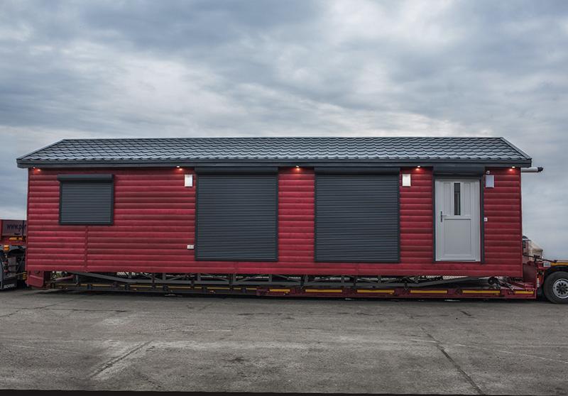 Czerwony dom szkieletowy z szarymi roletami zewnętrznymi i drzwiami, transportowany na niskopodwoziowej naczepie, widok z boku na tle pochmurnego nieba.