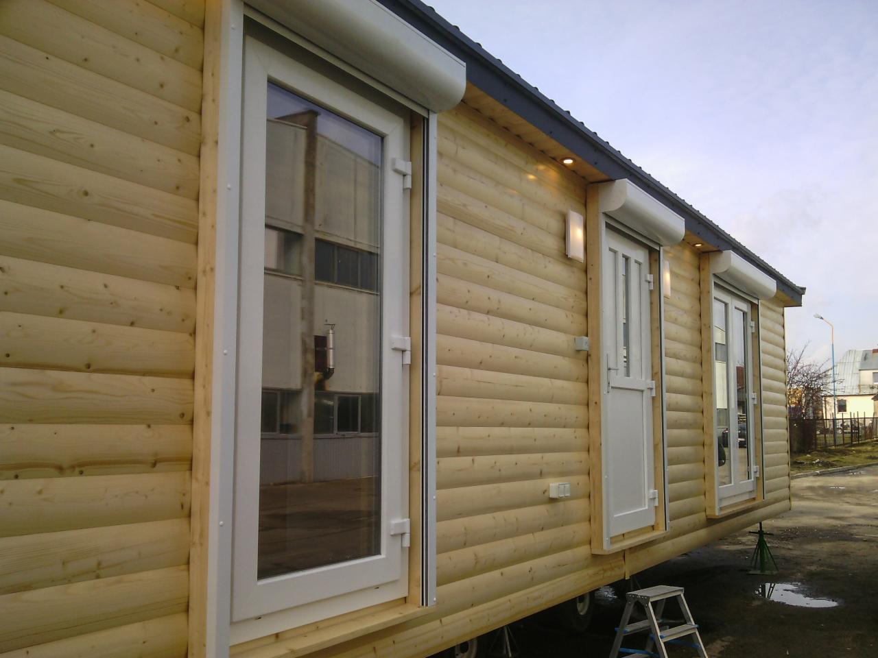 Zewnętrzny widok domu z drewnianych bali z trzema oknami i roletami zewnętrznymi, oświetleniem elewacyjnym i schodkami.