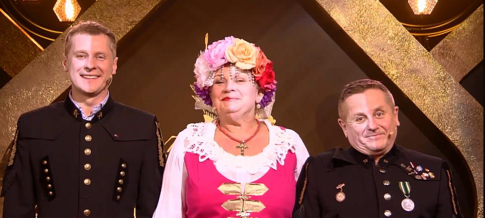 Trzy osoby w strojach ludowych lub galowych górniczych na tle dekoracji scenicznej z podświetleniem.