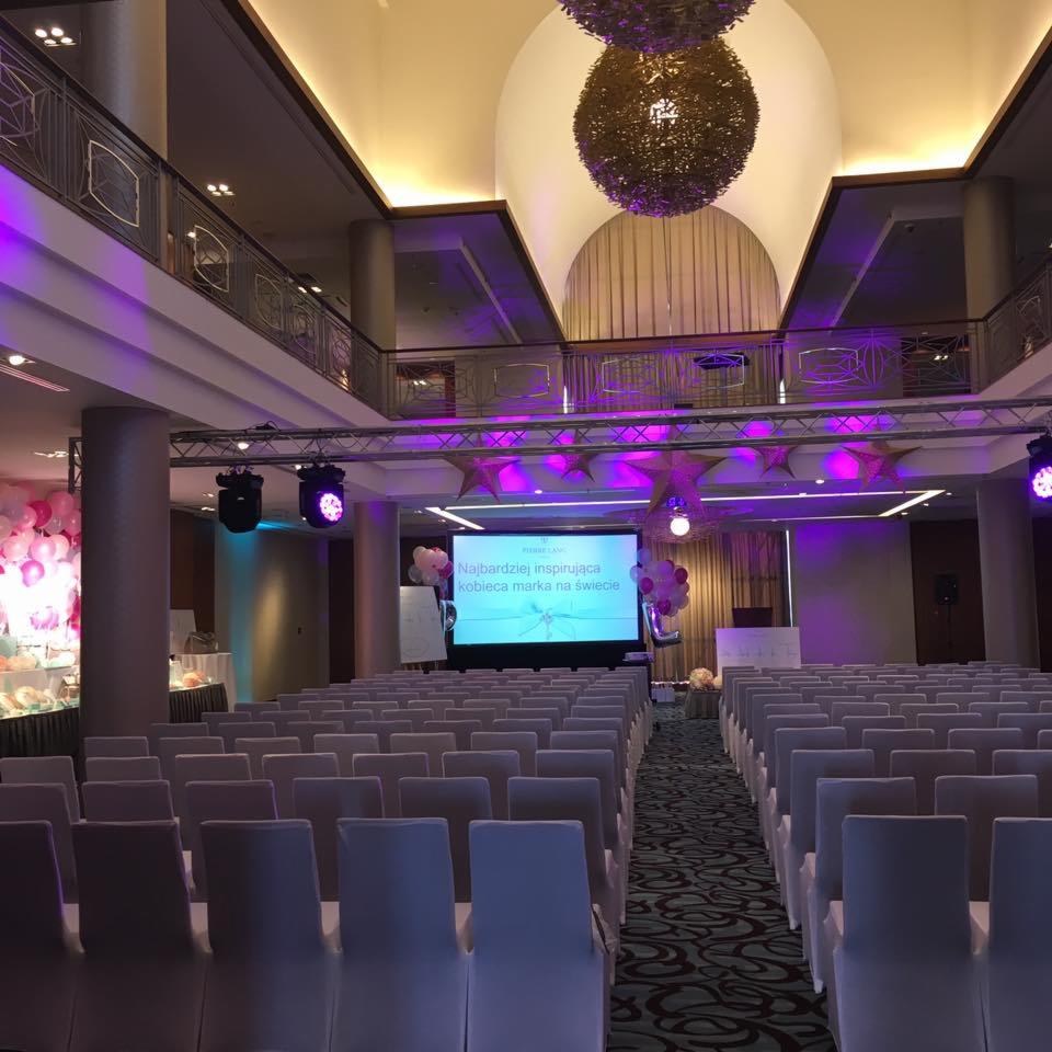 Sala konferencyjna przygotowana na event, z rzędami krzeseł, sceną z ekranem wyświetlającym napis i dekoracjami balonowymi, oświetlona fioletowym światłem.