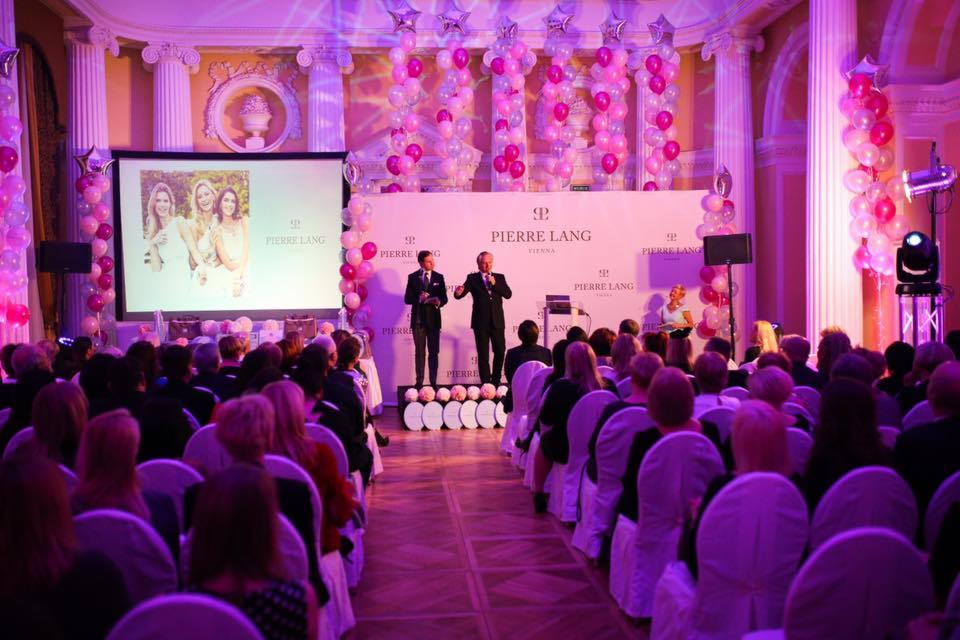 Konferencja firmy Pierre Lang w eleganckiej sali z kolumnami, różowymi balonami i oświetleniem, dwóch prelegentów na scenie przed publicznością.
