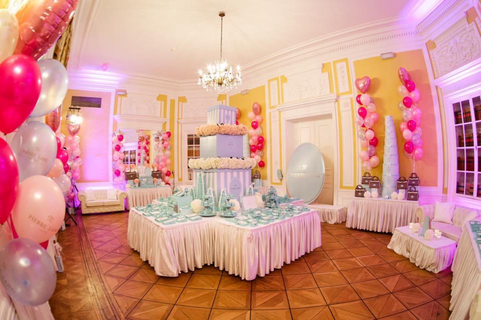 Elegancka sala udekorowana na przyjęcie z tortem z pudełek, balonami różowymi i białymi, oraz stołami z dekoracjami w odcieniach błękitu i bieli.