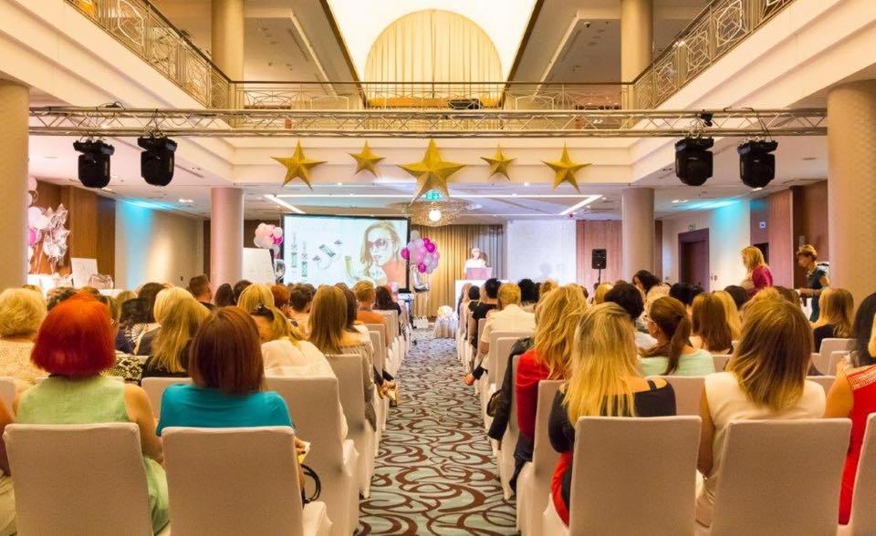 Sala konferencyjna z widokiem na scenę udekorowaną złotymi gwiazdami, rzędy krzeseł wypełnione publicznością, projektor wyświetla prezentację, oświetlenie sceniczne, kolumny i balkon w tle.