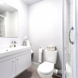 Unibase - Jasna łazienka z podwójną umywalką, kabiną prysznicową z chromowanymi uchwytami i toaletą z koszykiem na pokrywie.