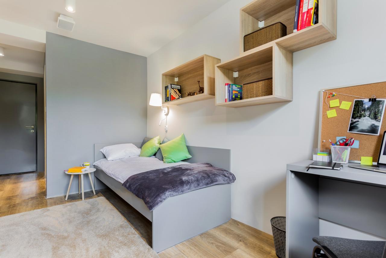 Pokój młodzieżowy z szarą sofą, drewnianymi półkami, biurkiem z korkową tablicą i jasnym dywanem.