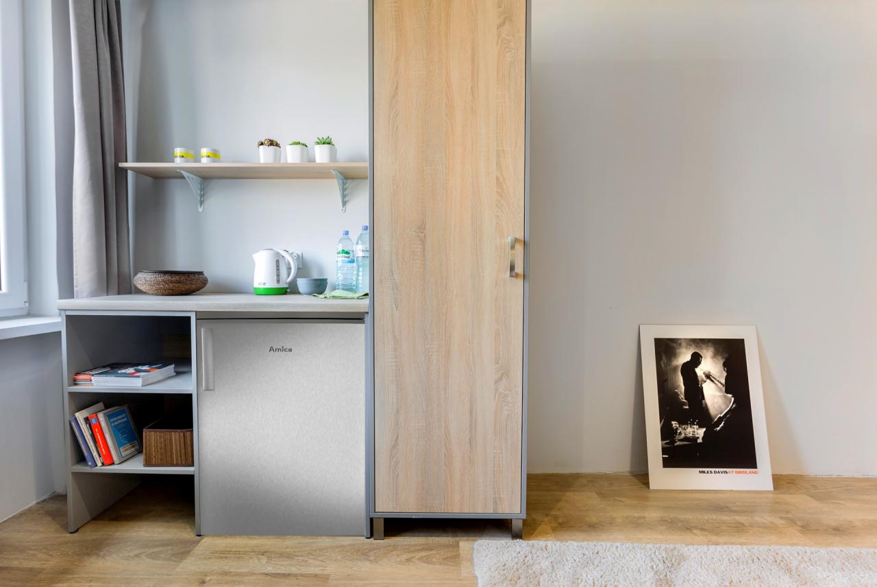 Wnętrze pokoju z aneksem kuchennym, widoczna szafka, lodówka Amica, półka z dekoracjami, czajnik elektryczny, woda butelkowana oraz plakat Milesa Davisa oparty o ścianę.