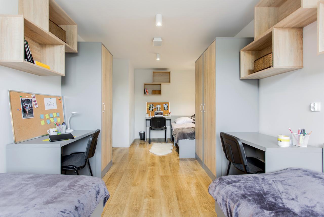 Wnętrze pokoju studenckiego z dwoma biurkami, łóżkami i szafami, utrzymane w szaro-beżowej kolorystyce, z tablicami korkowymi nad biurkami.