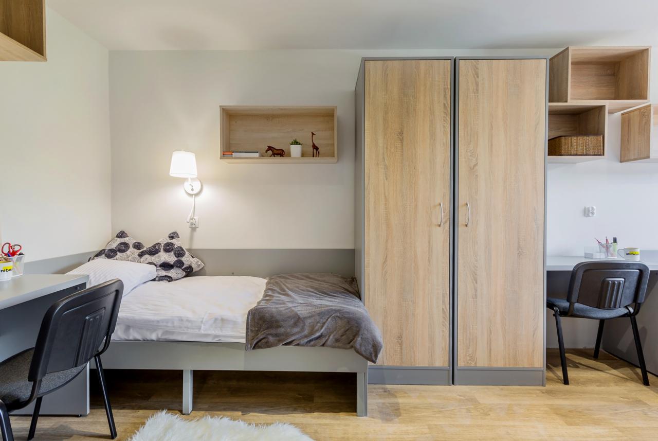 Pokój do wynajęcia: łóżko z szarą ramą, białą pościelą i szarym kocem, biurko z krzesłem, drewniana szafa, półki na ścianie z dekoracjami, lampka nocna i dywanik.