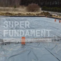 Przygotowany teren pod fundamenty z izolacją, zbrojeniem i pionowymi rurami kanalizacyjnymi. Widoczny teren budowy i częściowo wykończony dom.