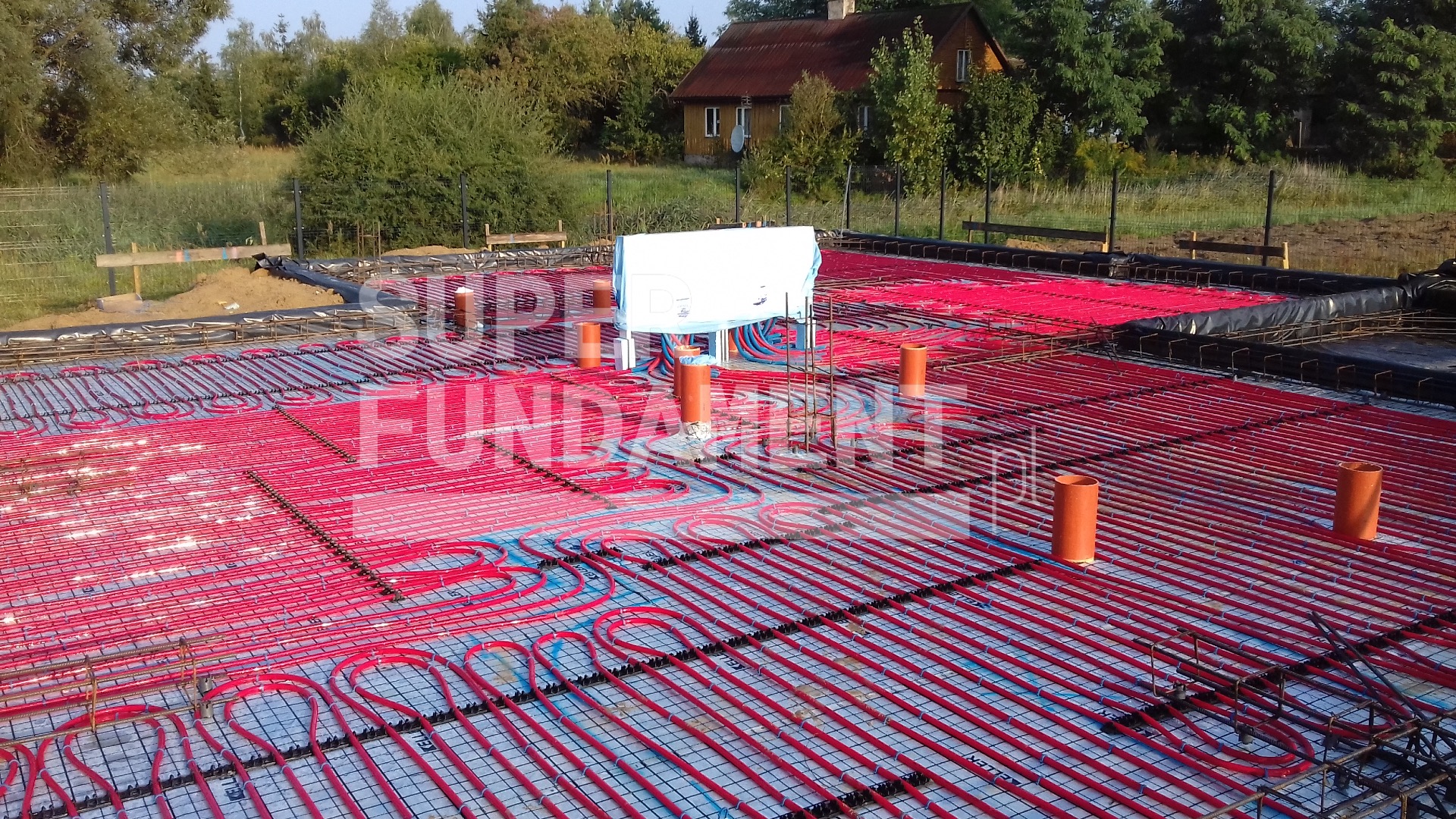 Fundamenty z ogrzewaniem podłogowym w trakcie budowy, rury grzewcze w kolorze czerwonym ułożone na izolacji, widoczne elementy zbrojenia i pionowe rury kanalizacyjne.