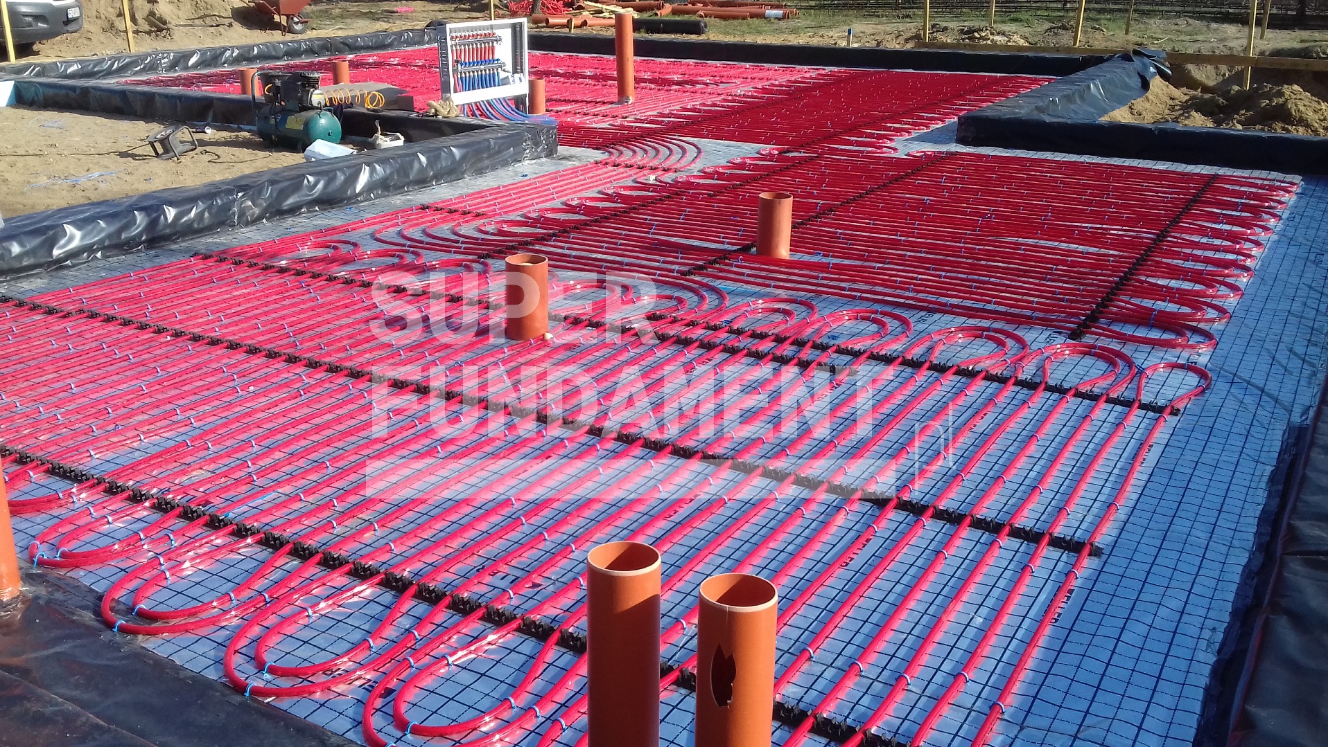 Instalacja ogrzewania podłogowego w fundamencie: czerwone rury ułożone na siatce, widoczne elementy instalacyjne i folia izolacyjna na obrzeżach.
