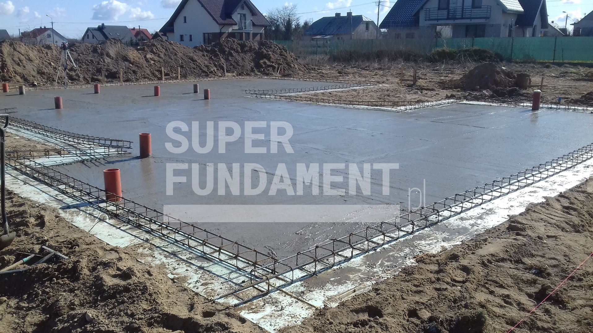 Wylane fundamenty z widocznym zbrojeniem i pionowymi rurami w gruncie. W tle domy jednorodzinne i hałda ziemi.