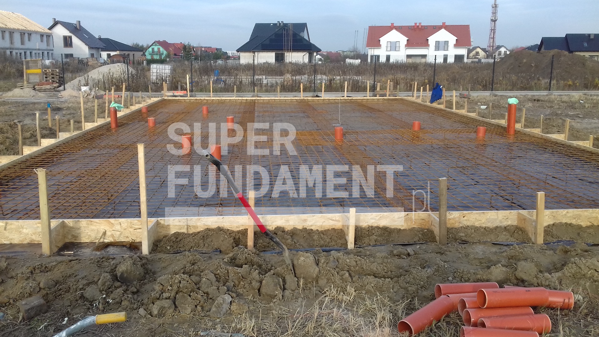 Zbrojenie fundamentów na budowie, widoczne szalunki, siatka zbrojeniowa, pionowe rury, narzędzia i ziemia. W tle domy jednorodzinne.