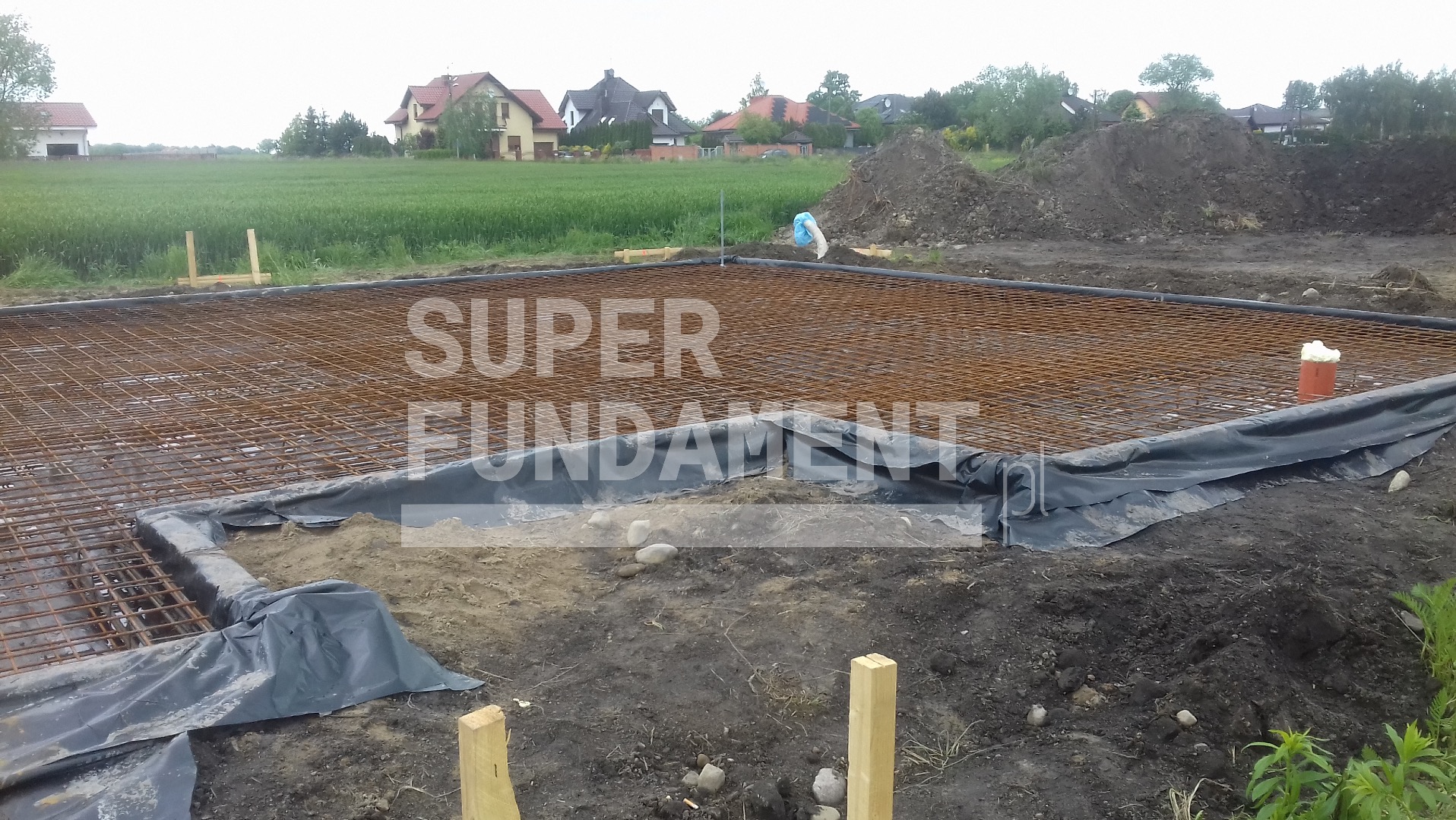 Zbrojenie fundamentów z folią hydroizolacyjną na terenie budowy, w tle zabudowa jednorodzinna i pole uprawne. Widoczne drewniane stemple.