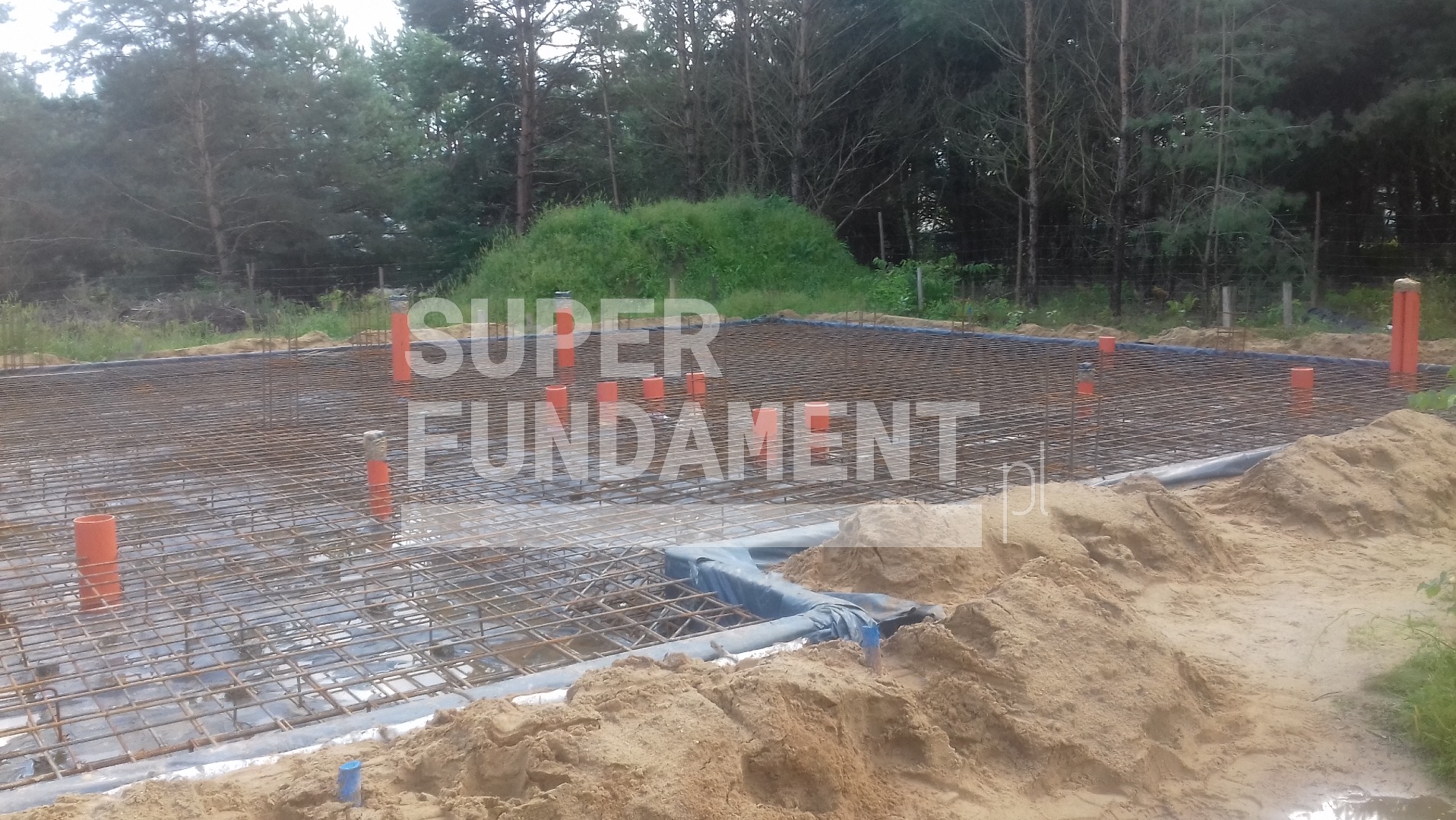 Zbrojenie fundamentów na folii, z wystającymi pionowo rurami i piaskiem na obrzeżach. Widoczne drzewa w tle.