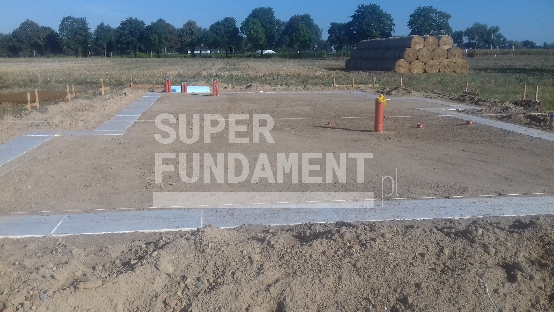 Wytyczone szalunki pod fundament z widocznymi rurami i izolacją, w tle sterta bel siana na zaoranym polu.