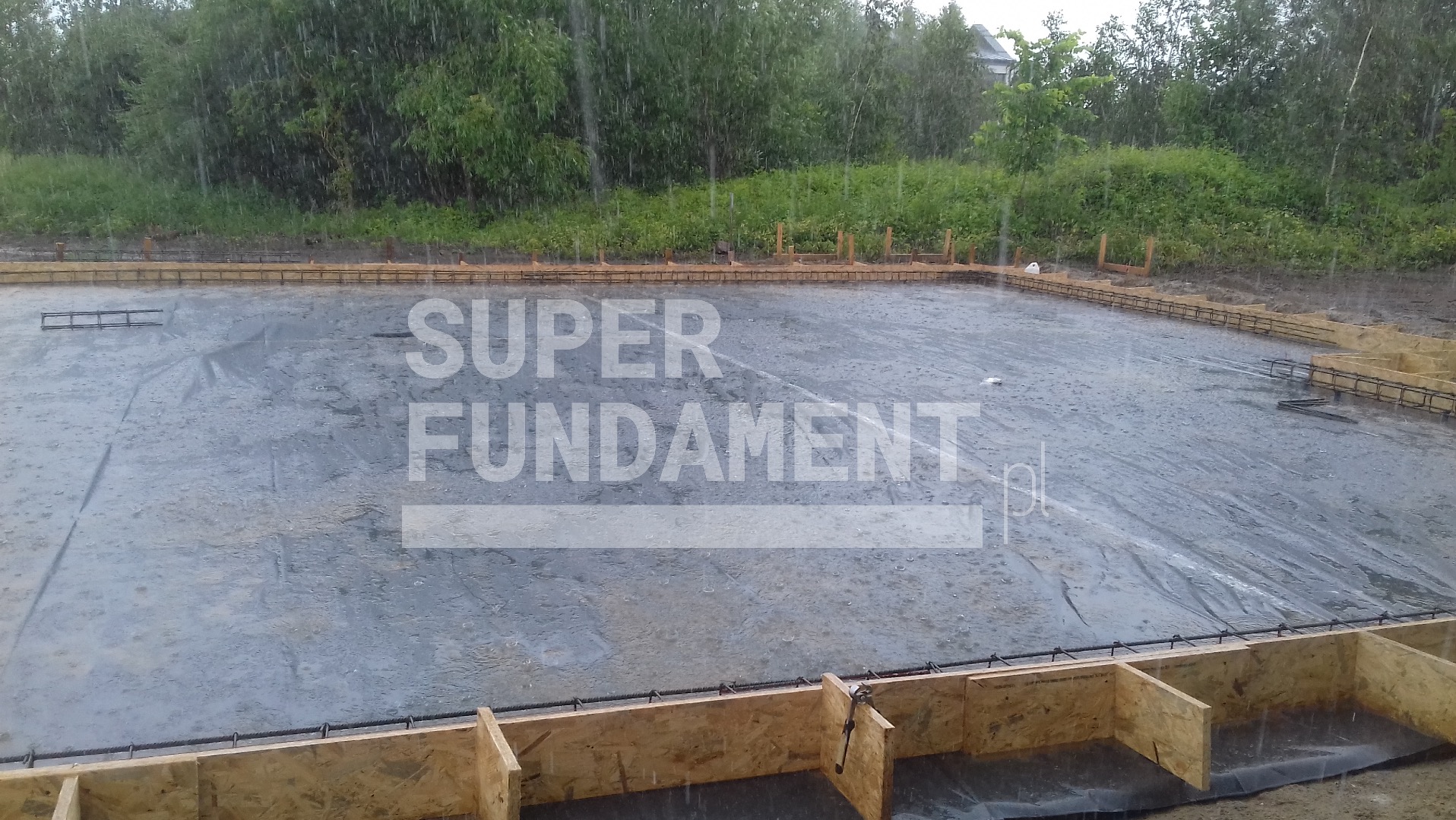 Deszcz na budowie fundamentów: szalunek z drewna, folia izolacyjna, zbrojenie. Widok na przygotowany teren pod wylewkę w deszczowy dzień.