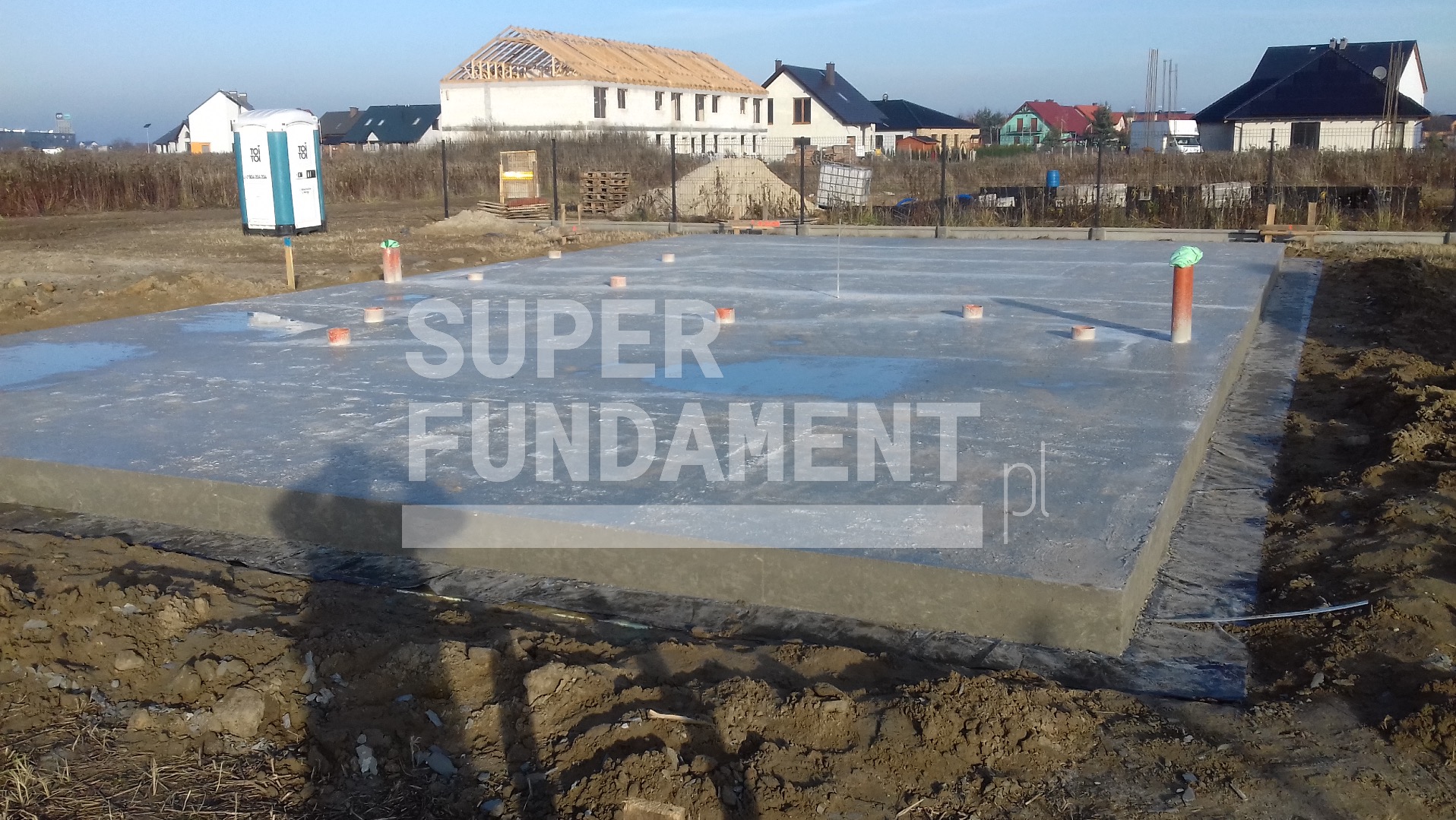 Wylany fundament z wystającymi rurami wentylacyjnymi na tle domów w budowie i przenośnej toalety. Widok z poziomu gruntu, wczesne popołudnie, słonecznie.