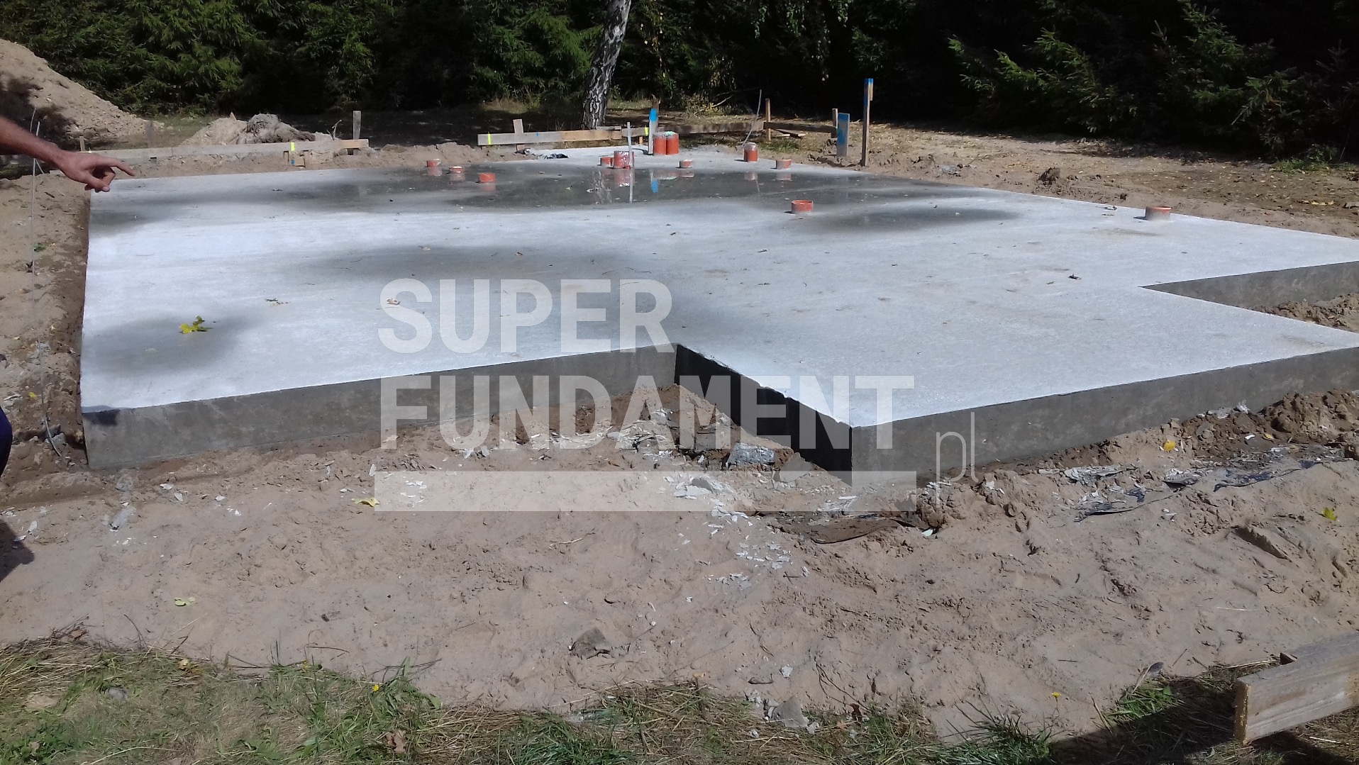 Wylany fundament z wystającymi rurami instalacyjnymi, otoczony ziemią. Widoczny fragment dłoni wskazującej na konstrukcję.