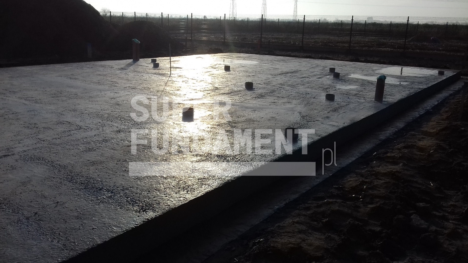 Zalana płyta fundamentowa z wystającymi rurami na budowie, widok z perspektywy, w tle ogrodzenie i hałda ziemi, oświetlone słońcem.