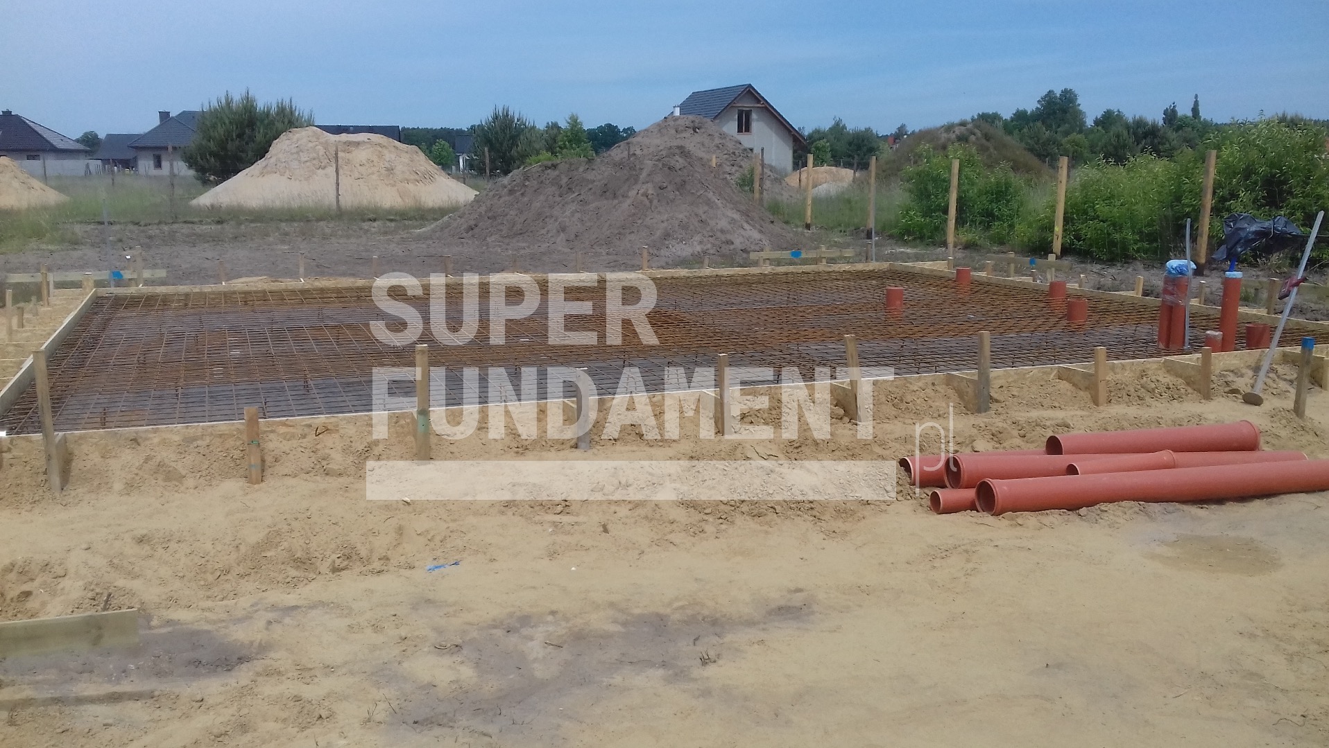 Przygotowanie zbrojenia i szalunku pod płytę fundamentową z widocznymi rurami kanalizacyjnymi i stertami ziemi w tle, w słoneczny dzień.