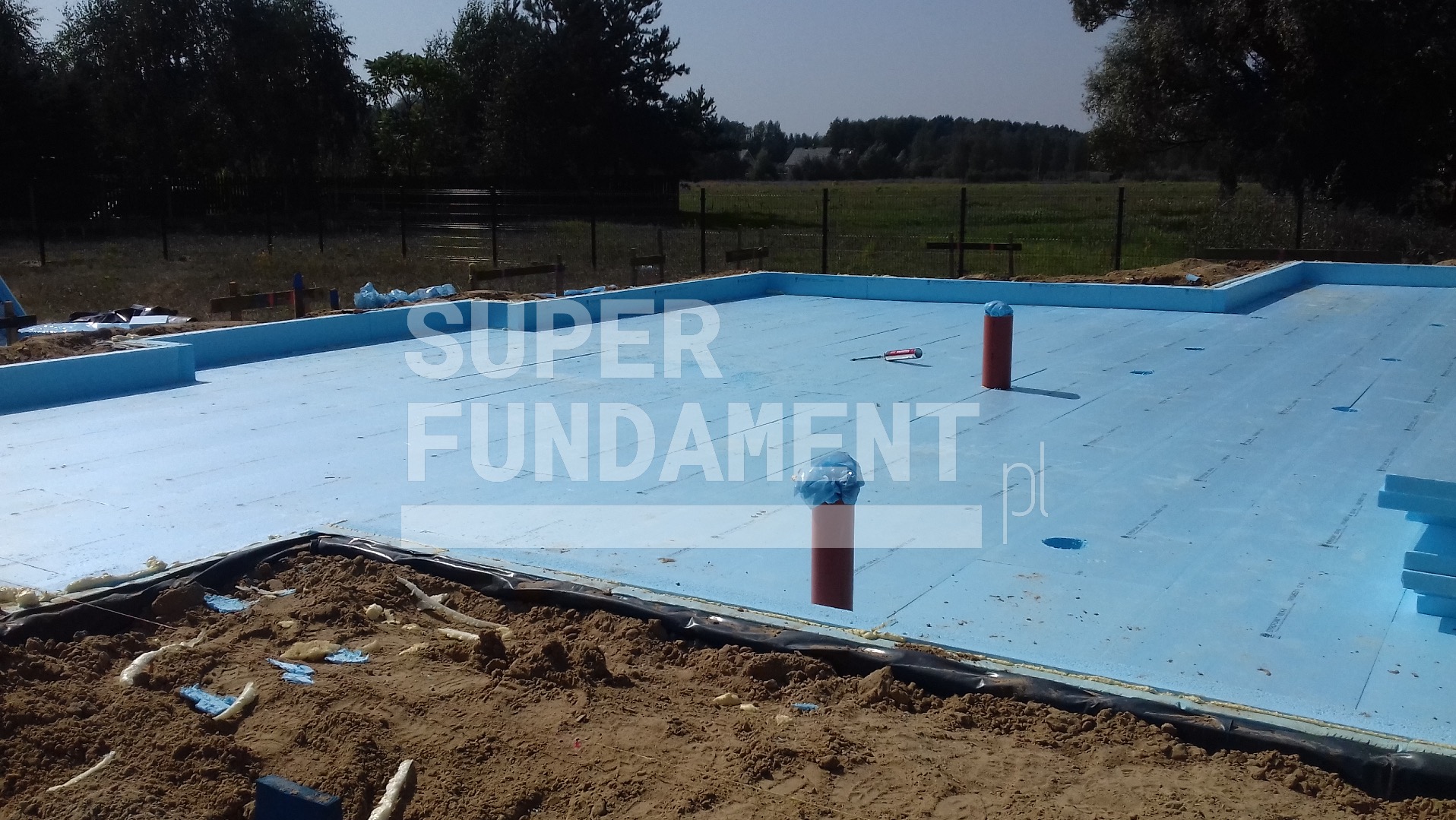 Ocieplone fundamenty z niebieskiego styropianu, widoczne rury instalacyjne zabezpieczone folią i narzędzie na powierzchni. Etap prac fundamentowych.