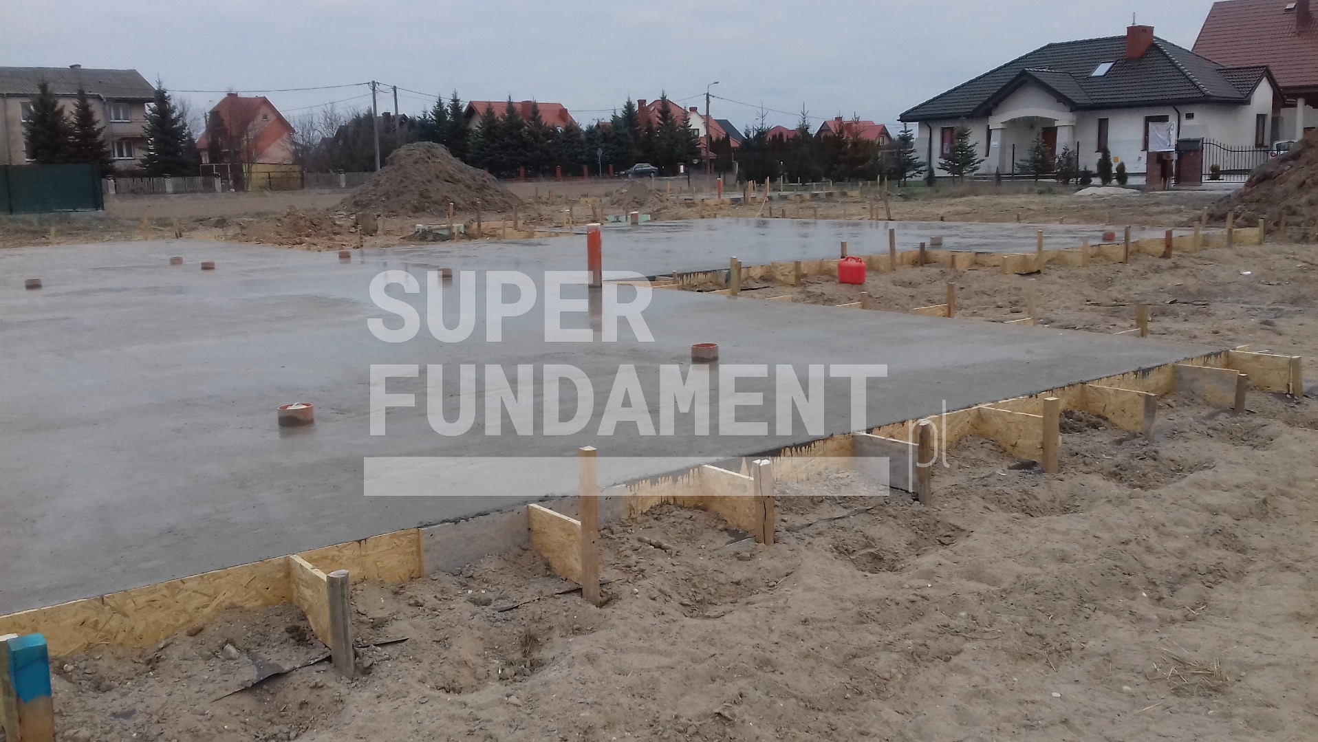 Zalany fundament z drewnianym szalunkiem na piaszczystym terenie, widok na domy w tle. Beton świeży, z widocznymi elementami poziomowania.