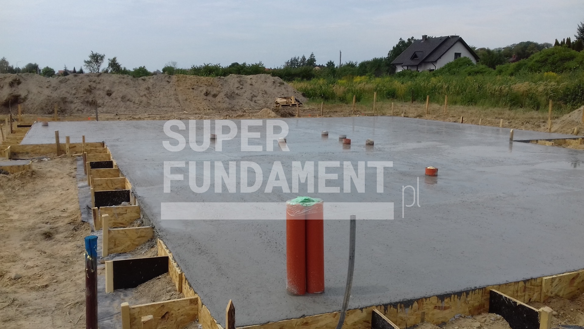 Zalane fundamenty z deskowaniem na obrzeżach. Widoczne pionowe rury instalacyjne i paleta cegieł w tle. Dom w oddali.