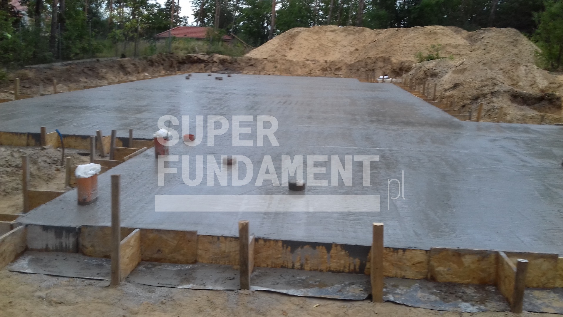 Wylana płyta fundamentowa w szalunku drewnianym, widoczne wiaderka i hałda piasku w tle. Surowy stan, przygotowanie do dalszych prac budowlanych.