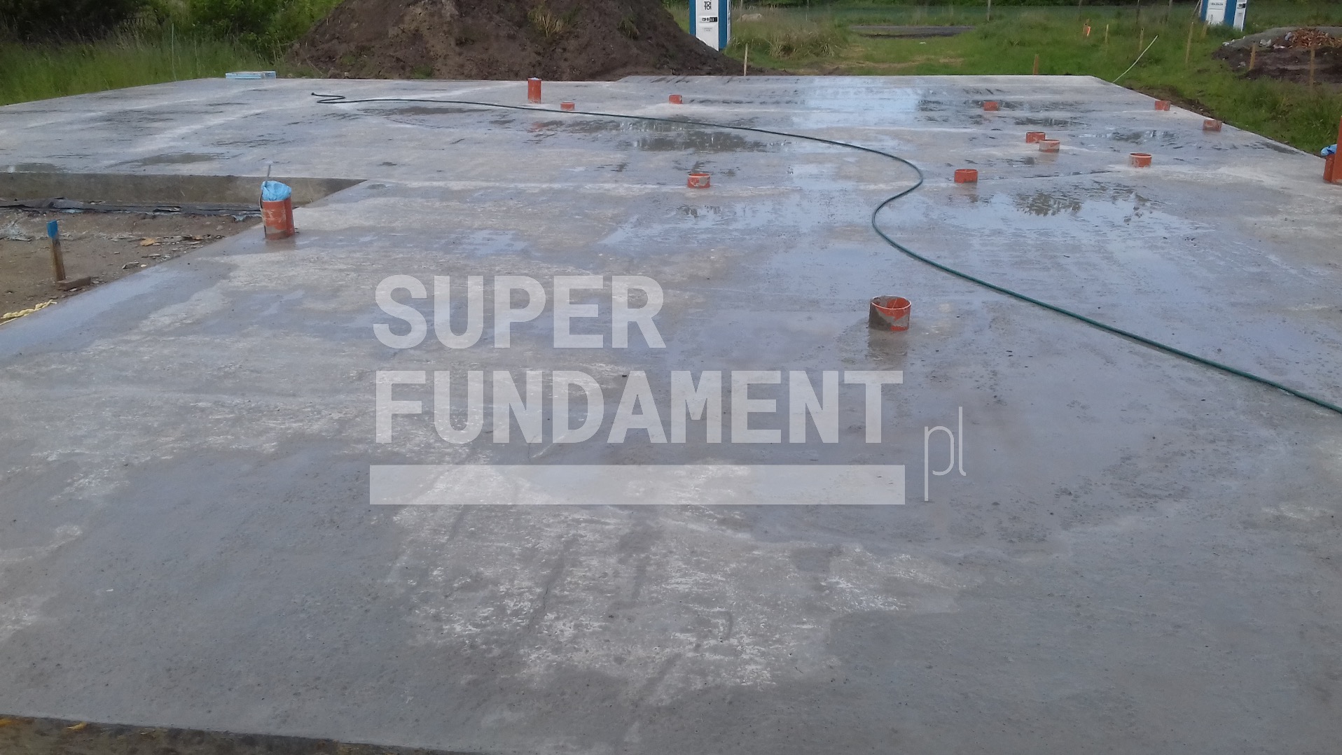 Wylana płyta fundamentowa z wystającymi rurami i zielonym wężem. Widoczne ślady po wodzie. W tle ziemia i roślinność.