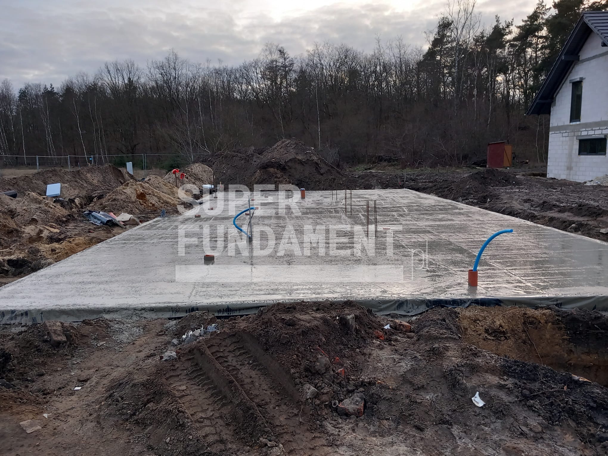 Wylana płyta fundamentowa z wystającymi rurami instalacyjnymi na tle ziemi i drzew, częściowo widoczny budynek w trakcie budowy.