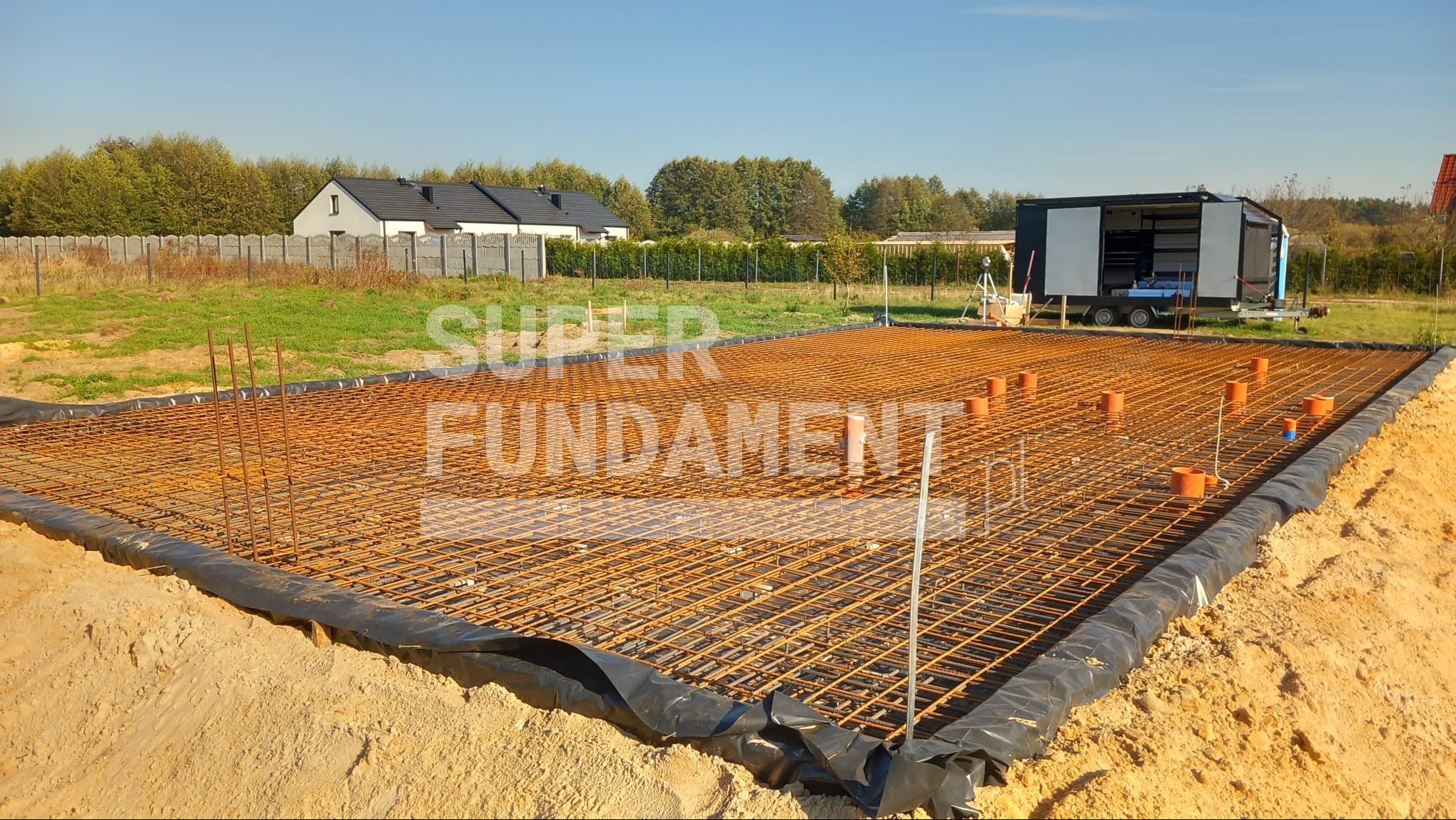 Zbrojenie płyty fundamentowej z widocznymi pionowymi prętami i pomarańczowymi rurami w gruncie. W tle kontener budowlany i dom jednorodzinny.