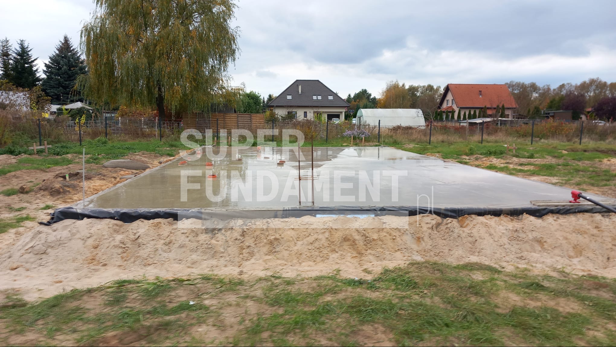 Wylane, mokre fundamenty płyty na piasku, zabezpieczone folią. W tle domy jednorodzinne i drzewa pod pochmurnym niebem.